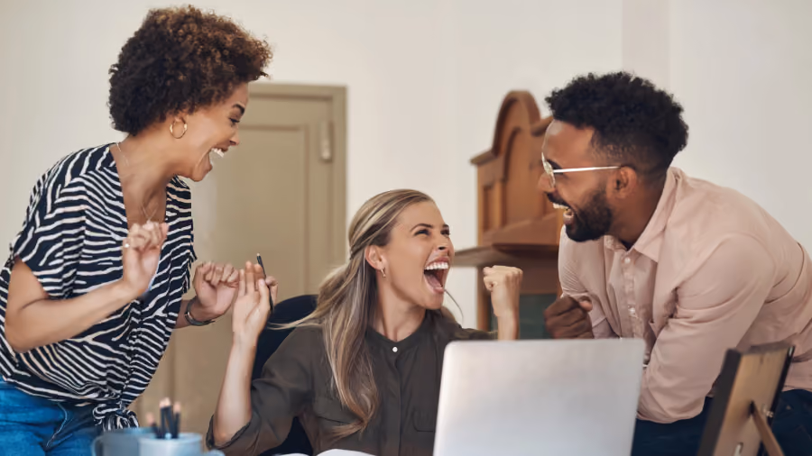 Foto de três colegas comemorando com os punhos erguidos e risadas em volta de um notebook sobre a mesa