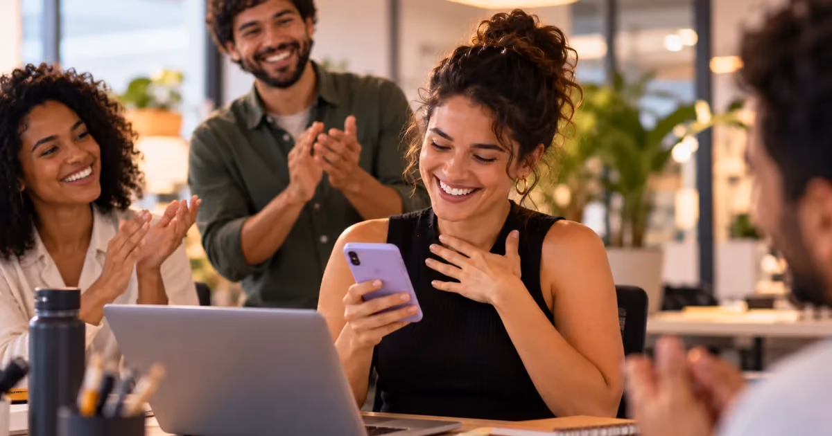 Mulher sorrindo ao olhar para o celular em uma mesa de escritório, enquanto três colegas ao redor aplaudem