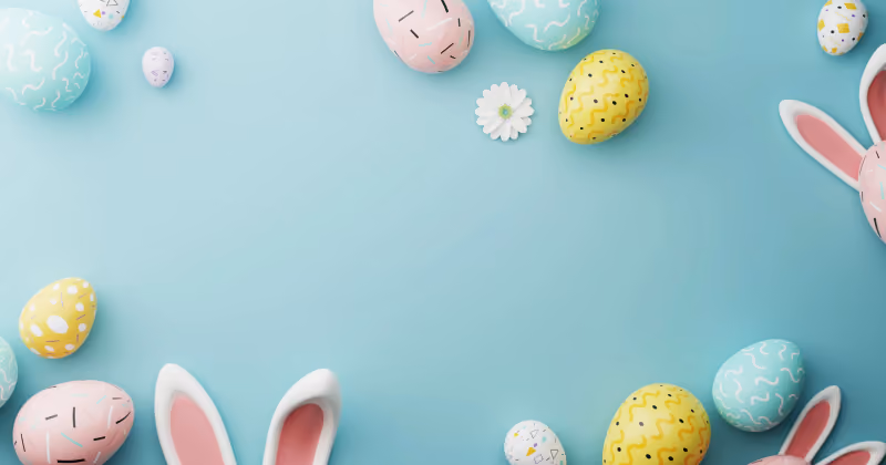 Foto de ovos de Páscoa em tons pastel, orelhas de coelho e uma margarida sobre fundo azul-claro, com espaço vazio ao centro