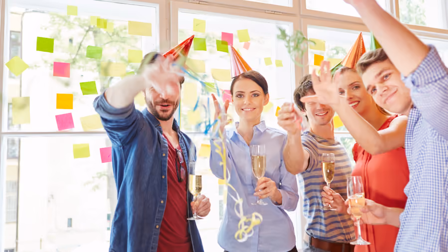 Foto de cinco pessoas com chapéus de festa e taças de espumante erguendo as mãos entre serpentinas, diante de parede com post-its