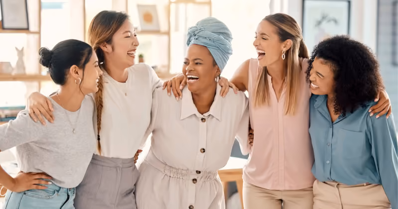 Foto de cinco mulheres abraçadas e rindo juntas em um escritório claro, ao lado de uma mesa de madeira
