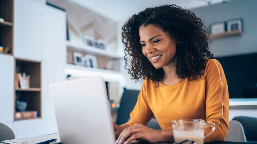 Foto de mulher de blusa amarela sorrindo enquanto digita no notebook em casa, com um copo de café ao lado