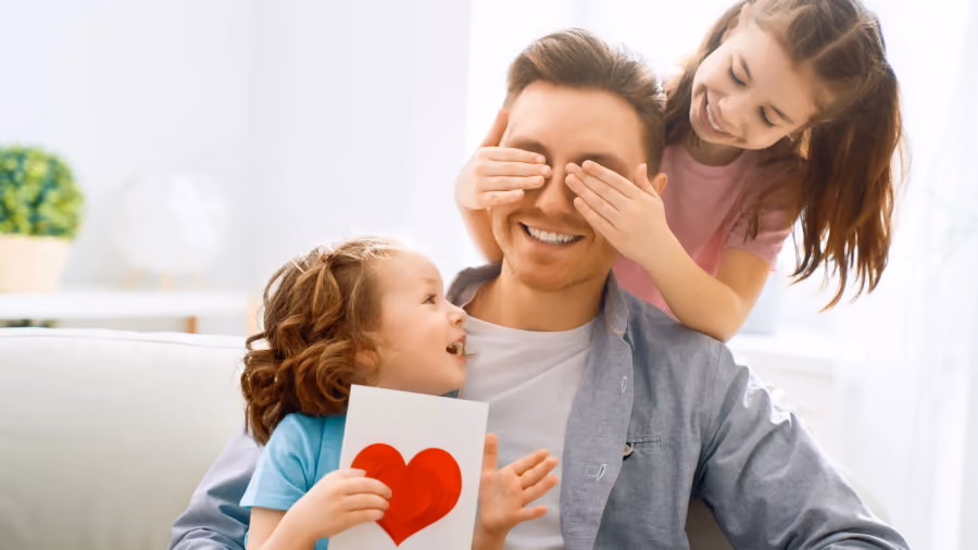 Foto de duas meninas com o pai no sofá: uma cobre os olhos dele por trás e a outra segura um cartão com coração vermelho