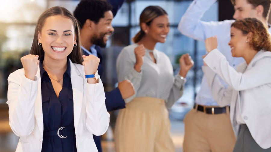 Foto de mulher de blazer branco sorrindo com os punhos cerrados em comemoração, com colegas celebrando desfocados ao fundo