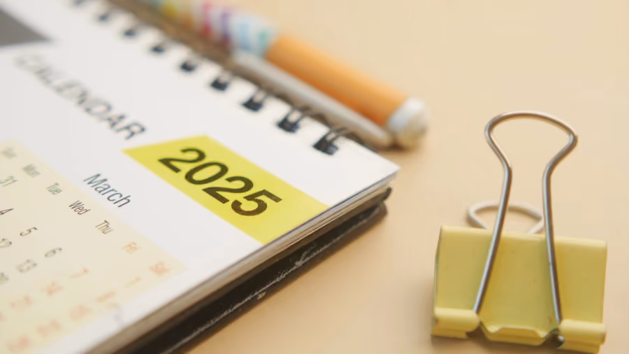Foto de calendário de mesa espiralado com marcação amarela do ano 2025, ao lado de um prendedor de papel amarelo e um lápis