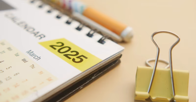 Foto de calendário de mesa espiralado com marcação amarela do ano 2025, ao lado de um prendedor de papel amarelo e um lápis