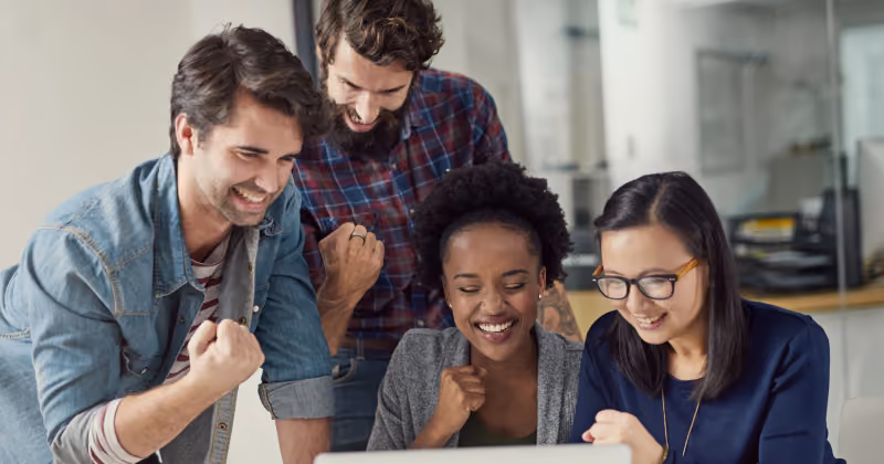 Foto de quatro colegas reunidos em volta de um notebook, sorrindo e comemorando com os punhos cerrados