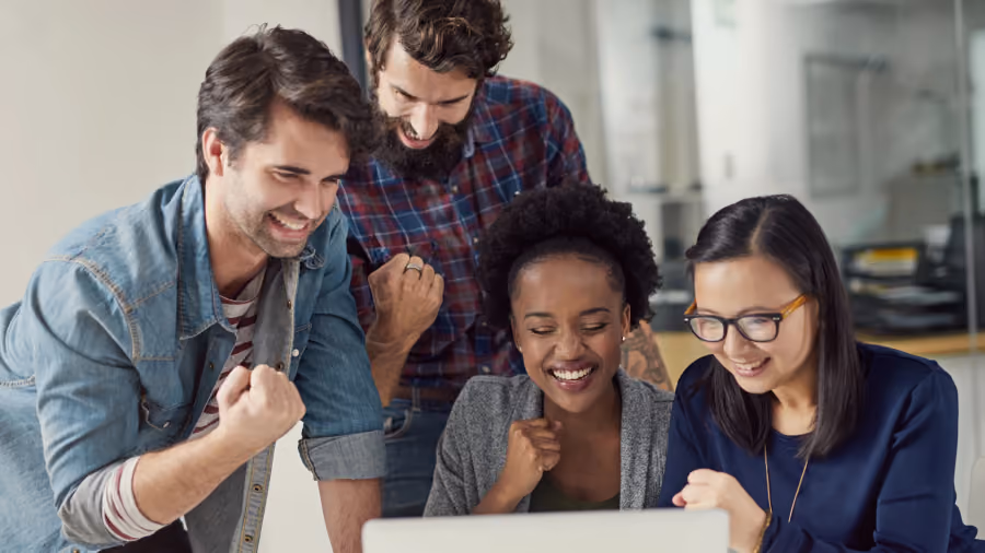 Foto de equipe de quatro pessoas olhando a tela de um notebook e vibrando com o resultado, punhos erguidos e sorrisos