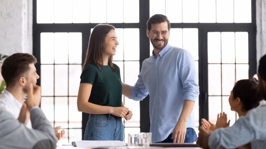 Foto de uma mulher e um homem em pé numa sala de reunião enquanto colegas sentados à mesa aplaudem