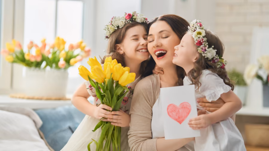 Foto de uma mãe abraçada por duas filhas com coroas de flores, segurando tulipas amarelas e um cartão com coração desenhado