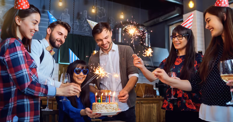Foto de seis pessoas com chapéus de festa em volta de um bolo com velas, segurando estrelinhas acesas e taças