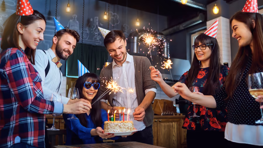 Foto de seis pessoas com chapéus de festa em volta de um bolo com velas, segurando estrelinhas acesas e taças
