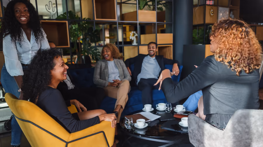 Foto de seis colegas rindo e conversando em poltronas de um lounge de escritório, com xícaras de café na mesa de centro