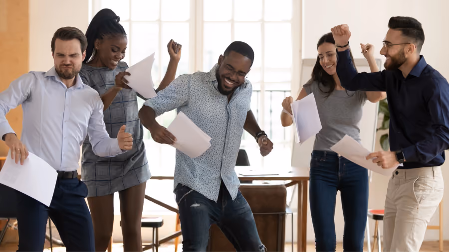 Foto de cinco pessoas comemorando de pé no escritório, com papéis nas mãos e punhos erguidos