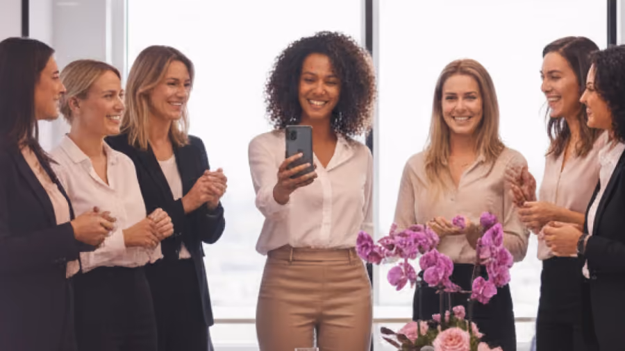 Foto de sete mulheres sorrindo e aplaudindo num escritório, uma delas com o celular na mão, e orquídeas em primeiro plano