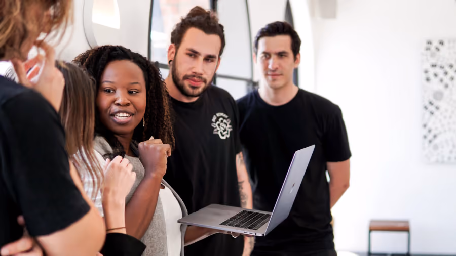 Foto de um grupo de jovens vestidos de preto em volta de um notebook, com uma mulher ao centro cerrando o punho e sorrindo