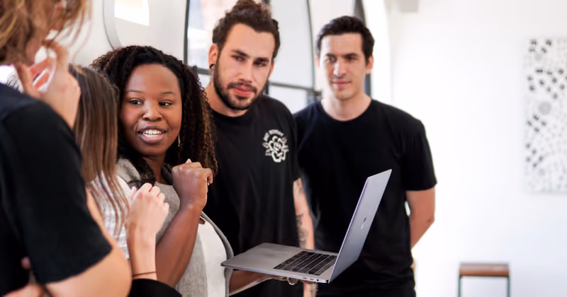Foto de um grupo de jovens vestidos de preto em volta de um notebook, com uma mulher ao centro cerrando o punho e sorrindo