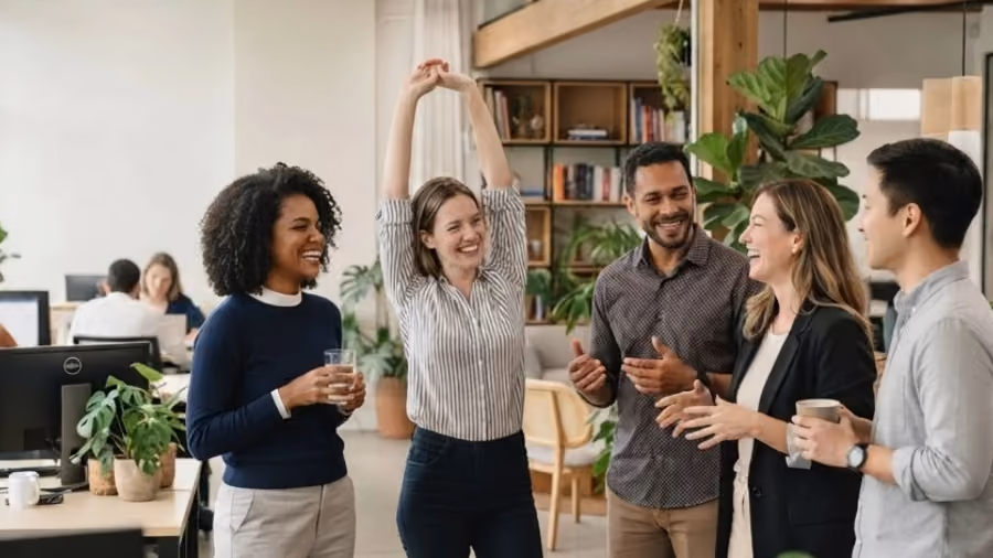Foto de cinco colegas conversando e rindo em escritório com plantas, uma delas com os braços erguidos em alongamento