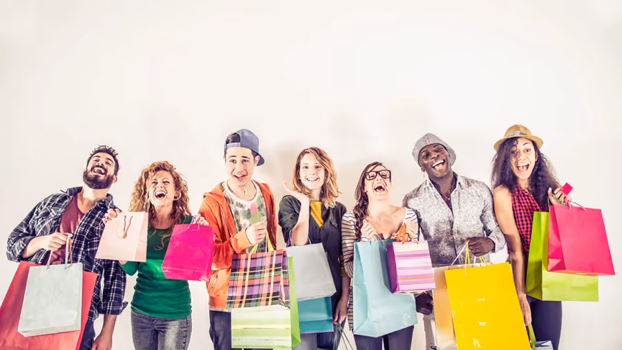 Foto de sete jovens sorrindo lado a lado contra parede branca, segurando várias sacolas de compras coloridas e um cartão rosa