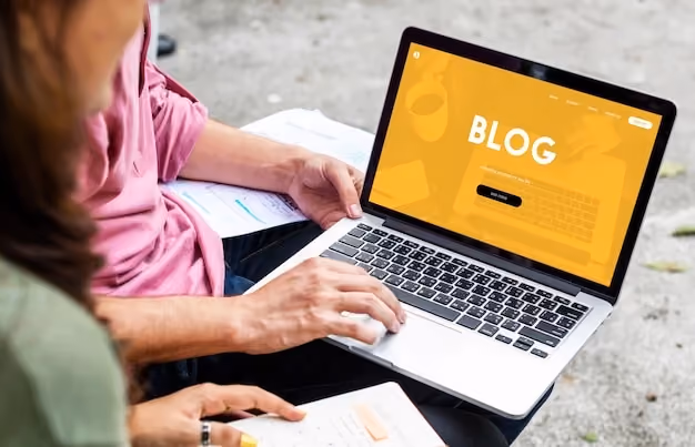 Foto por cima do ombro de pessoa sentada com notebook no colo, exibindo site amarelo com a palavra blog, ao lado de anotações