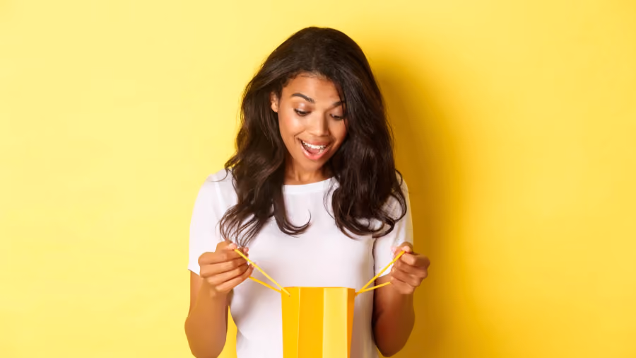 Foto de jovem de camiseta branca sobre fundo amarelo, sorrindo ao abrir e olhar dentro de uma sacola de presente