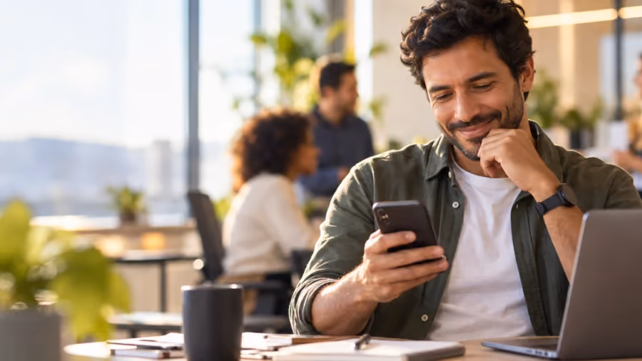 Foto de homem sorrindo enquanto olha o celular, sentado a uma mesa de escritório com notebook, com colegas ao fundo