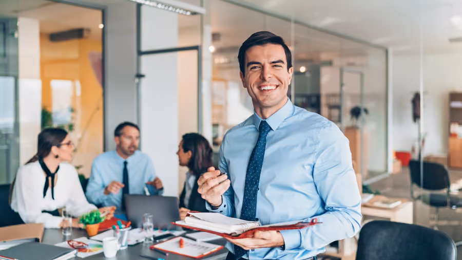 Foto de homem de camisa azul e gravata sorrindo com uma pasta vermelha aberta nas mãos, com colegas reunidos ao fundo