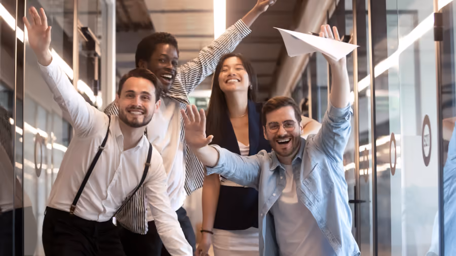 Foto de quatro colegas comemorando num corredor de escritório com os braços erguidos, um deles segurando um avião de papel