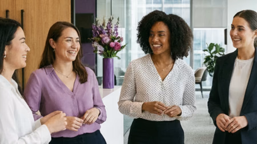 Foto de quatro mulheres conversando e sorrindo em um escritório, ao lado de um vaso com flores roxas