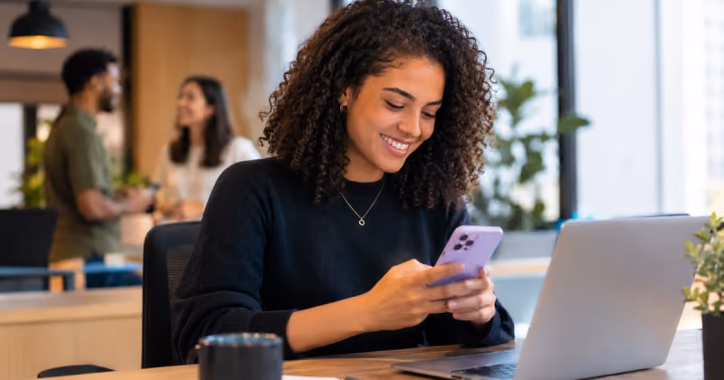Mulher sorrindo ao olhar para o celular em uma mesa de escritório, com um notebook ao lado e dois colegas conversando ao fundo