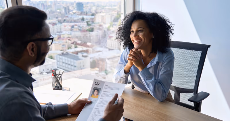 Foto de entrevista de emprego junto a uma janela, com o recrutador segurando um currículo e a candidata sorrindo