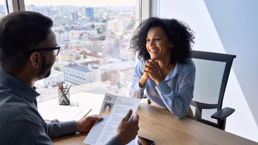 Foto de entrevista de emprego junto a uma janela, com o recrutador segurando um currículo e a candidata sorrindo