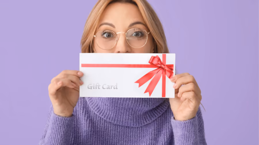 Foto de mulher de óculos segurando diante do rosto um envelope branco com laço vermelho e a inscrição Gift Card