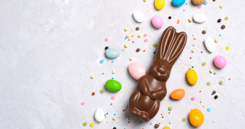Foto vista de cima de um coelho de chocolate cercado por ovinhos coloridos e confeitos sobre superfície cinza