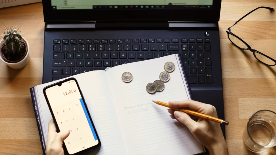 Foto vista de cima de mesa com notebook, caderno aberto com moedas, celular na calculadora e mão segurando lápis