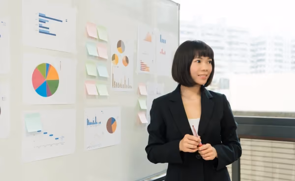 Foto de mulher de blazer preto ao lado de um quadro branco coberto de gráficos impressos e post-its coloridos