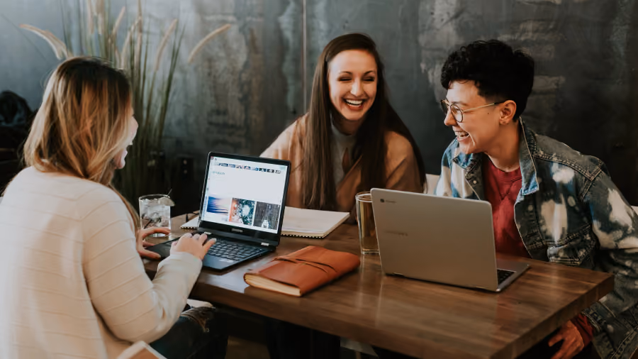 Foto de três mulheres conversando e rindo ao redor de uma mesa de madeira com dois notebooks e cadernos abertos