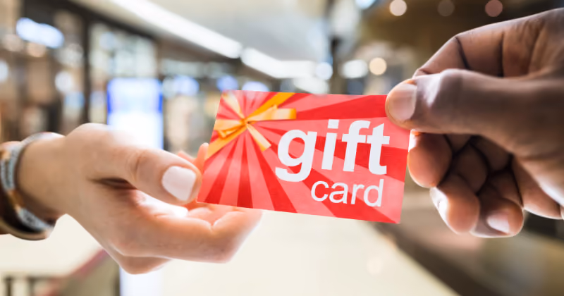 Foto de duas mãos passando um cartão vermelho com a inscrição gift card, tendo um shopping desfocado ao fundo