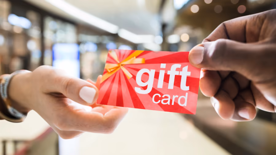 Foto de duas mãos passando um cartão vermelho com a inscrição gift card, tendo um shopping desfocado ao fundo